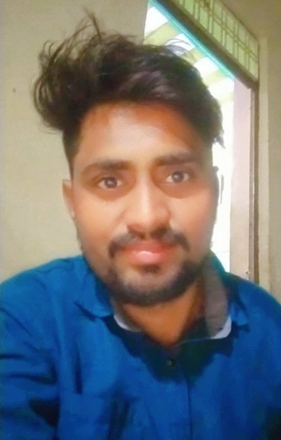 A young man with dark hair and a beard taking a selfie indoors, wearing a blue shirt.