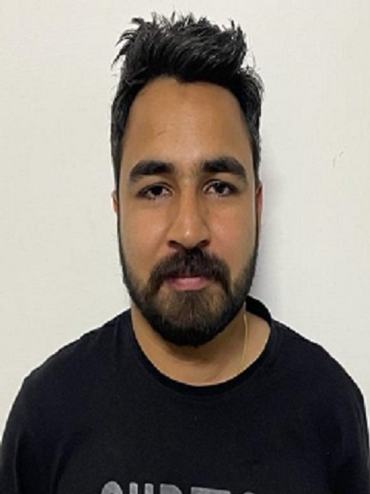 A man with dark hair and a beard wearing a black T-shirt standing against a plain white wall.
