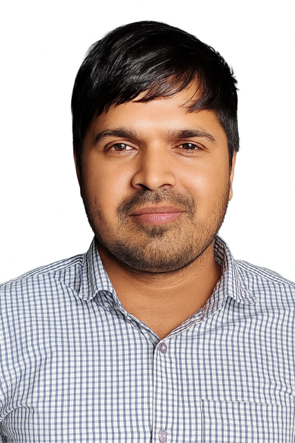 A man with medium skin tone, black hair, and a beard, wearing a light blue checkered button-up shirt, looking directly at the camera with a neutral expression.