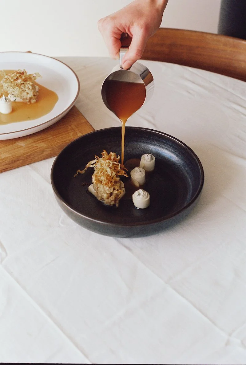 Persoon giet saus over een gerecht op een zwarte bord.