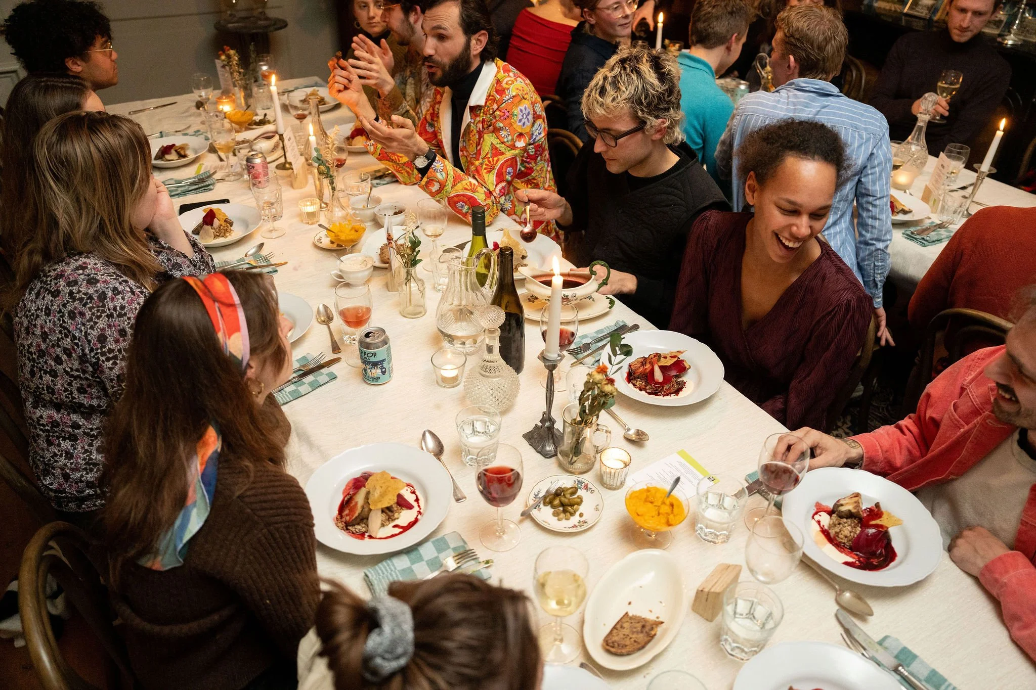 Deelnemers van de SFYN academie 2025 zitten aan een lange tafel tijdens een diner, lachen en praten, met diverse gerechten en drankjes.