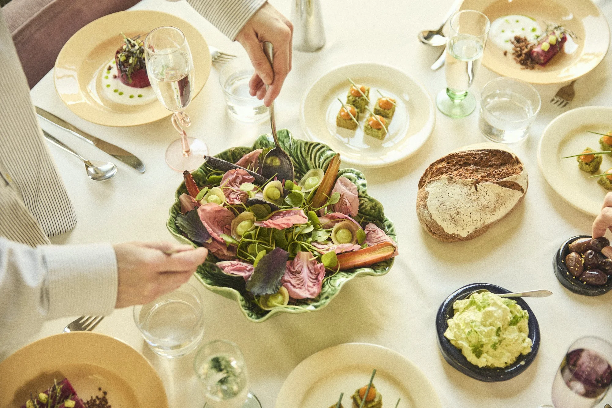 Een tafel met verschillende gerechten, waaronder een kleurrijke salade met groene en roze bladeren, kleine stukjes brood met toppings, een brood met een zachte, lichte korst, en diverse kleine schaaltjes met andere hapjes en drankjes, inclusief glaasjes met water en wat wijn.