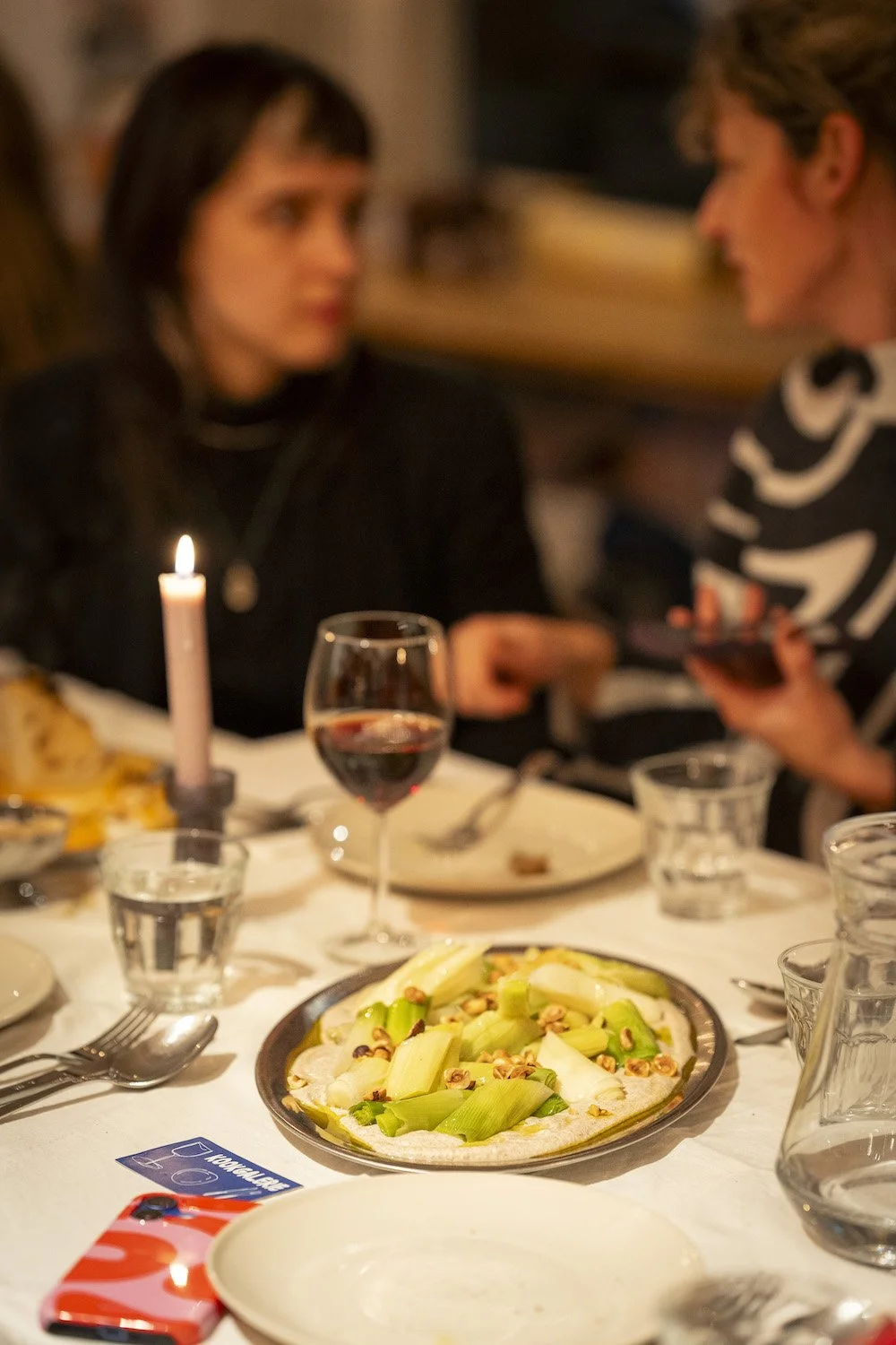 Een diner tafelschikking met witte tafelkleed, een bord met prei, een glas rode wijn, een glas water, een kaars, en twee mensen die met elkaar praten.