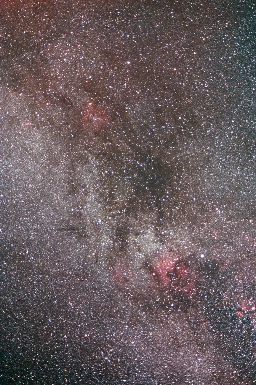 A detailed photograph of the dense star field of the Milky Way galaxy, showing countless stars, dark interstellar dust clouds, and some pink-colored emission nebulae.