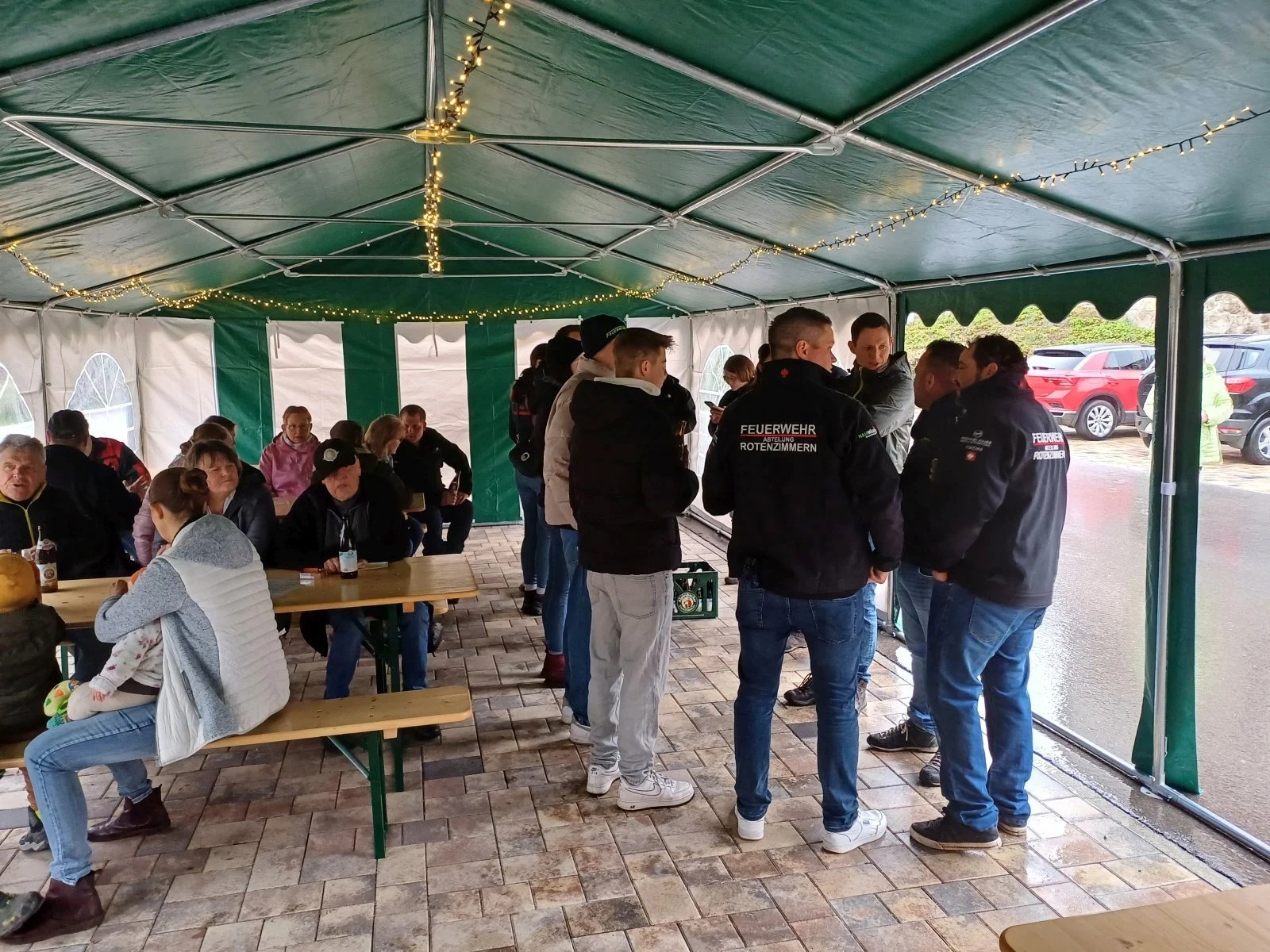 Menschen versammelt in einer Zelthalle, einige tragen Feuerwehruniform, sitzen an Tischen oder stehen im Gespräch, bei schlechtem Wetter im Freien.