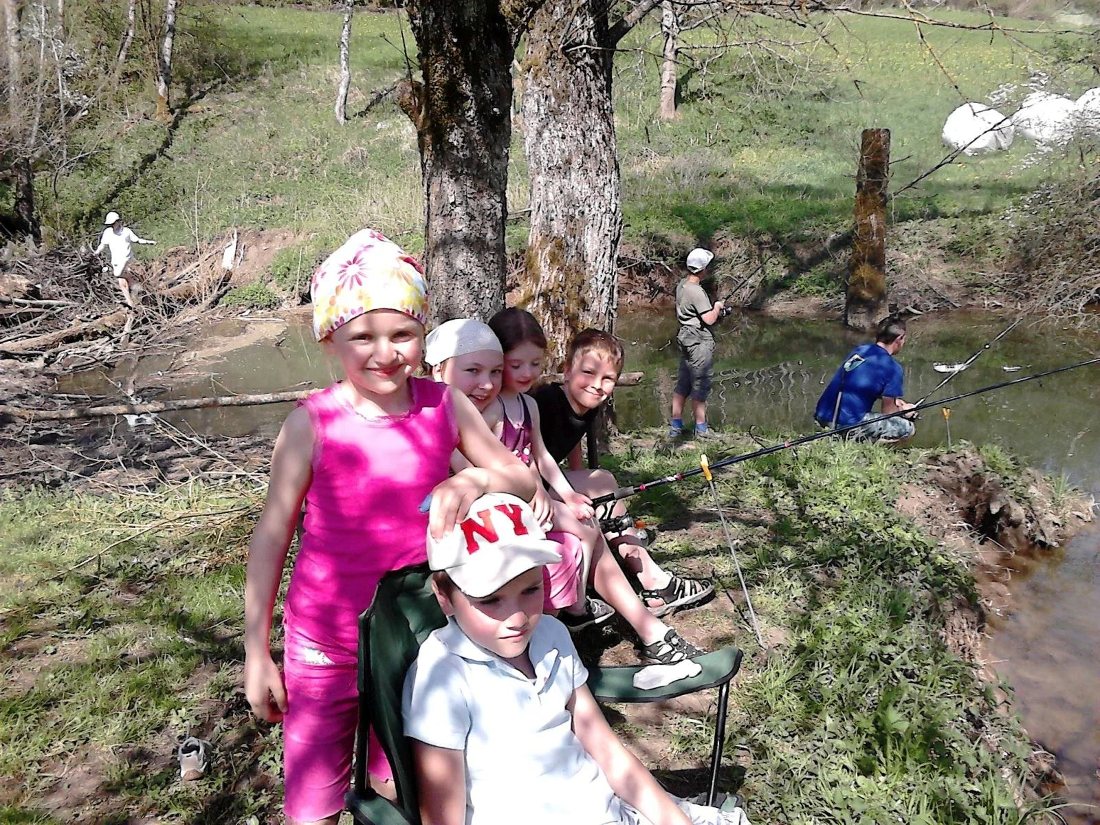 Eine Gruppe von Kindern sitzt und steht am Ufer eines Flusses, während einige von ihnen angeln. Es ist ein sonniger Tag im Freien, umgeben von Bäumen und Natur.