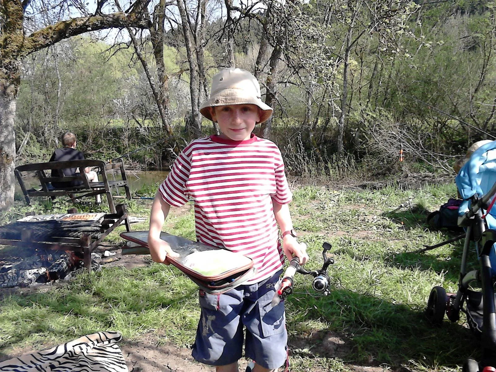 Ein Junge mit Sonnenhut und gestreiftem T-Shirt steht mit einem Fahrrad in einem Waldgebiet, während im Hintergrund eine Person am Ufer eines Flusses sitzt und grillt.