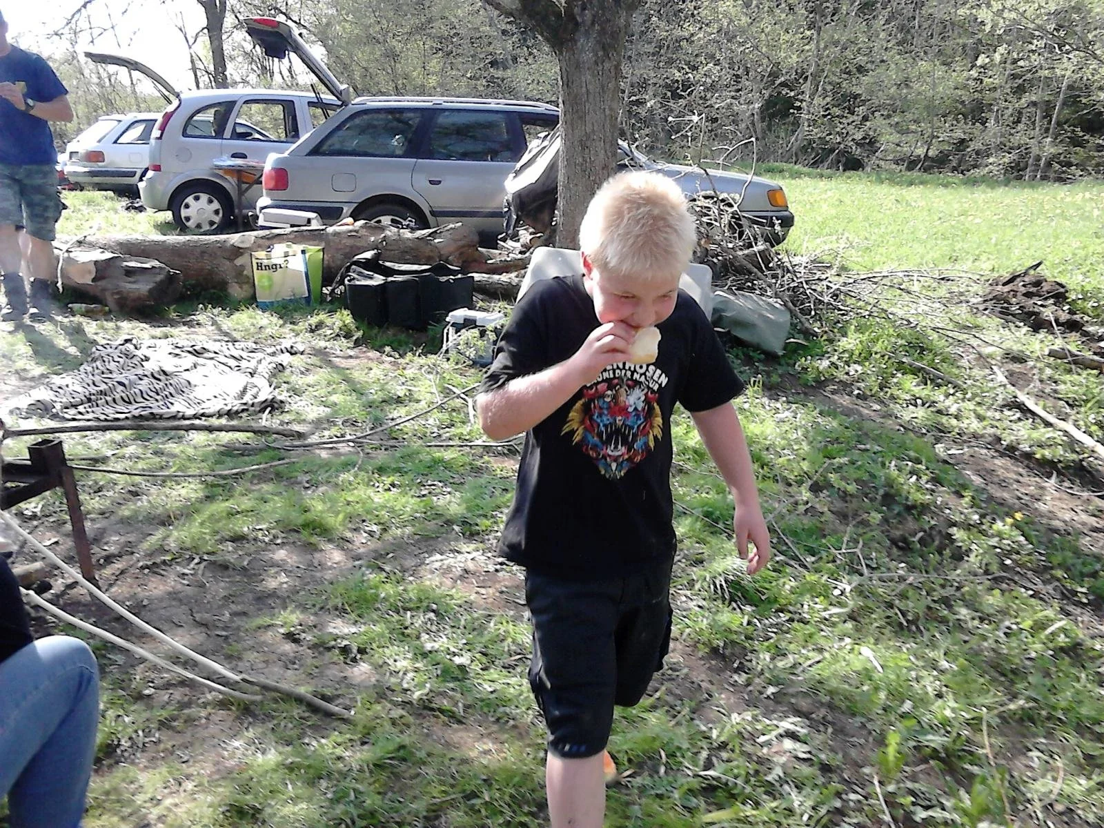 Ein junger Junge mit blondem Haar isst ein Stück Brot im Freien, im Hintergrund sind Fahrzeuge, Bäume und andere Personen zu sehen.
