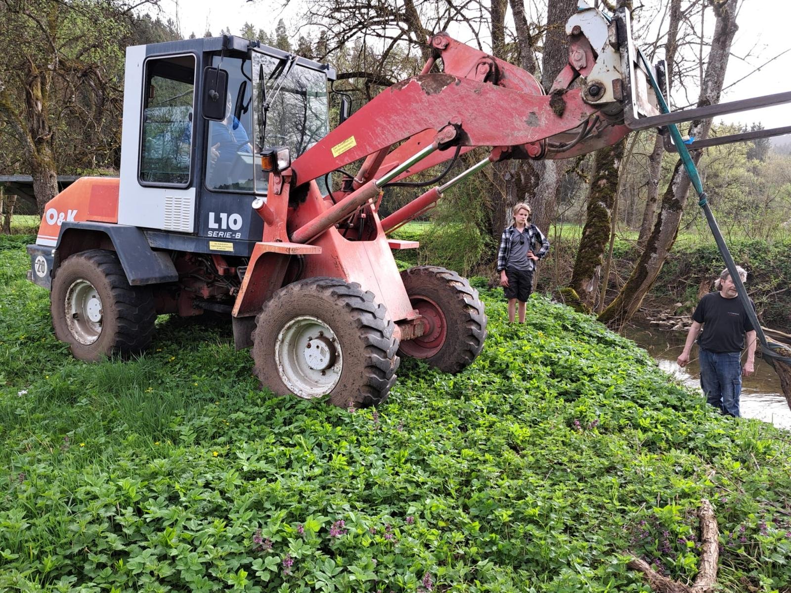 Ein roter Bagger steht auf einem grünen, bewachsenen Ufer neben einem Fluss. Zwei Menschen, ein Mann und eine Frau, stehen in der Nähe, die Frau auf dem Hügel und der Mann im Wasser, während sie das Geschehen beobachten.