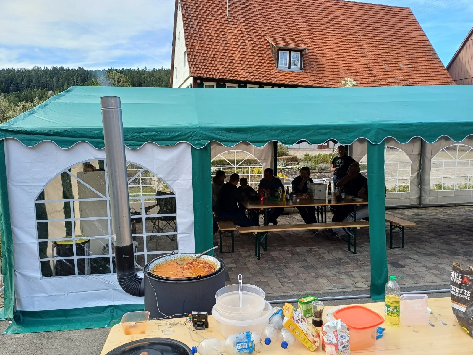 Blick auf eine grüne Partyzelt mit mehreren Personen, die darunter sitzen, einige stehen, bei einer Feier im Freien. Im Vordergrund ist ein Tisch mit Suppe, Getränken und Essensresten. Im Hintergrund Häuser und Himmel zu sehen.