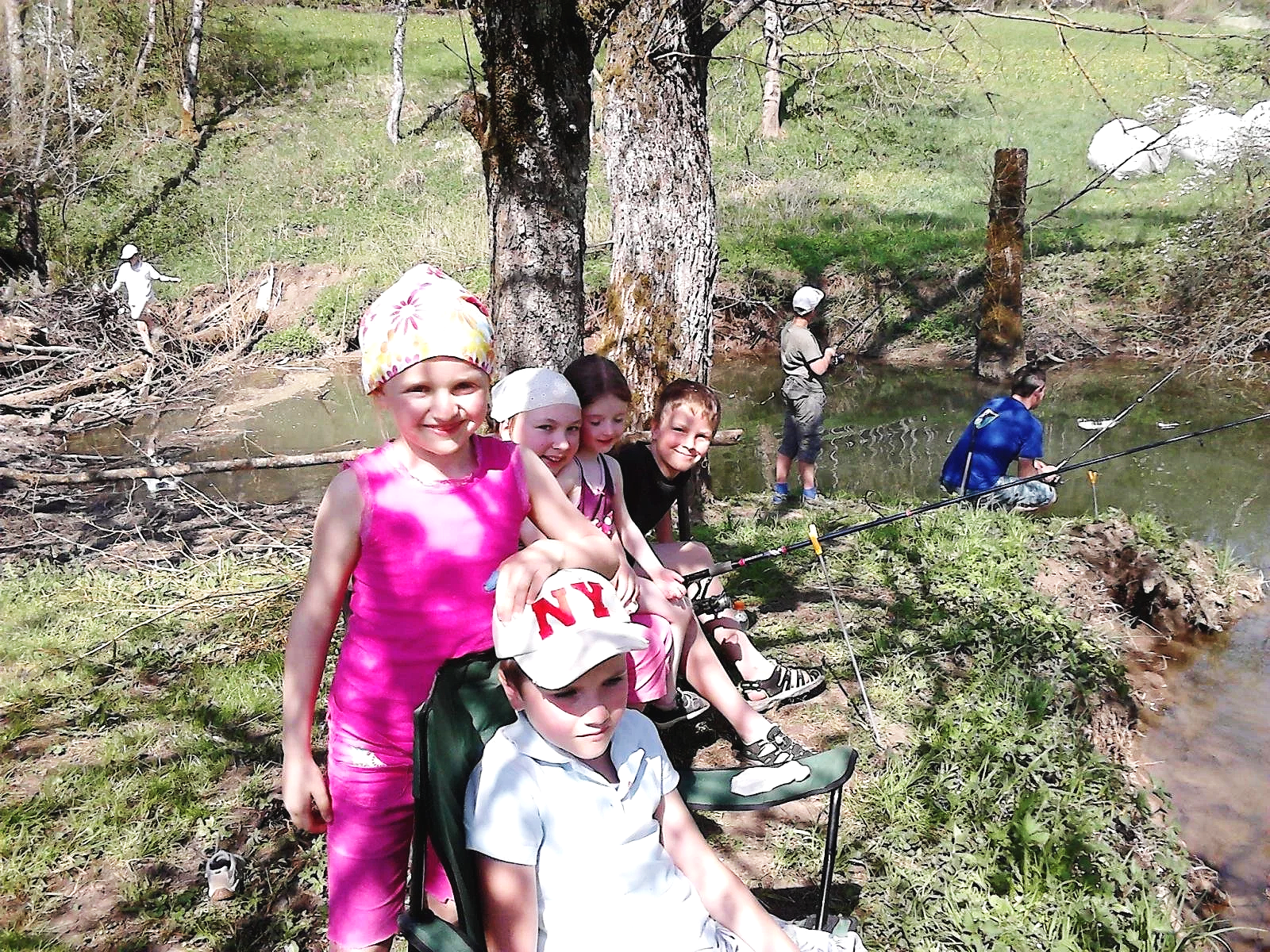 Gruppe von Kindern, die am Fluss angeln, sitzen und stehen am Ufer, einige mit Angeln, im Hintergrund Bäume und Wasser.