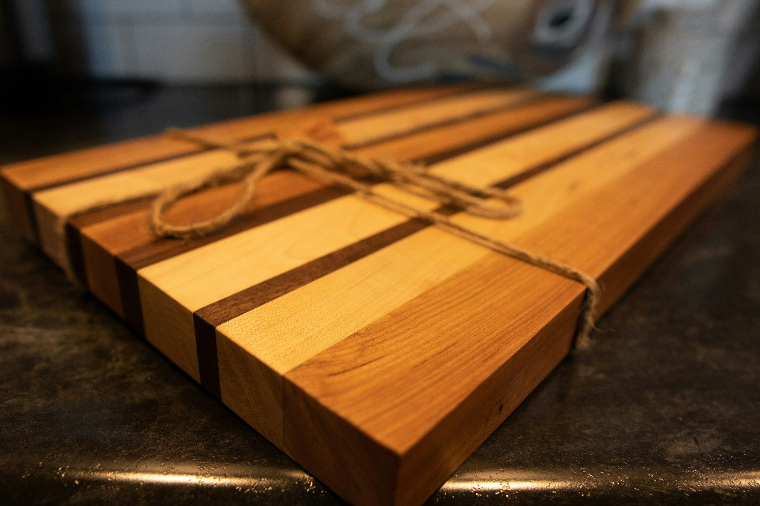 A wooden cutting board with alternating light and dark wood stripes, tied with a piece of twine, placed on a dark countertop.