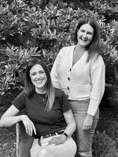 Two women smiling outdoors, one sitting and one standing, with bushes in the background.