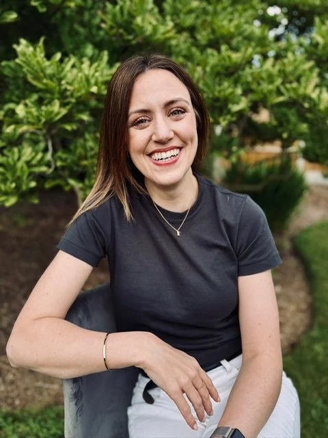 A young woman with shoulder-length brown hair, wearing a black t-shirt, sitting outdoors on a bench, smiling at the camera with greenery in the background.
