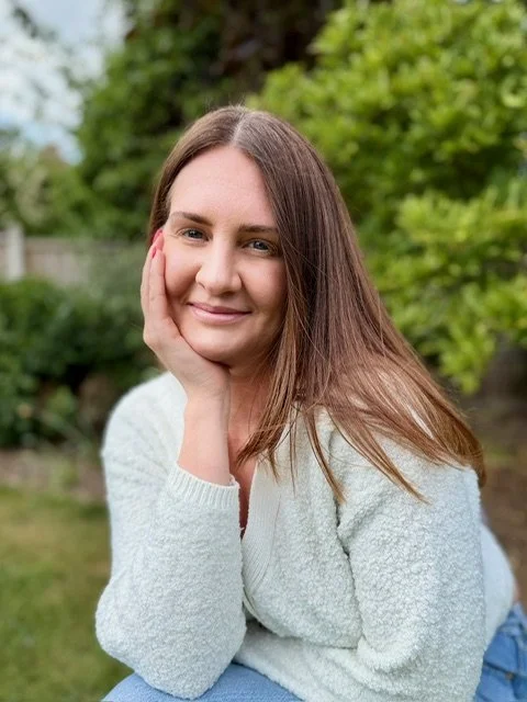 A young woman with long brown hair smiling in an outdoor setting with trees and greenery in the background.