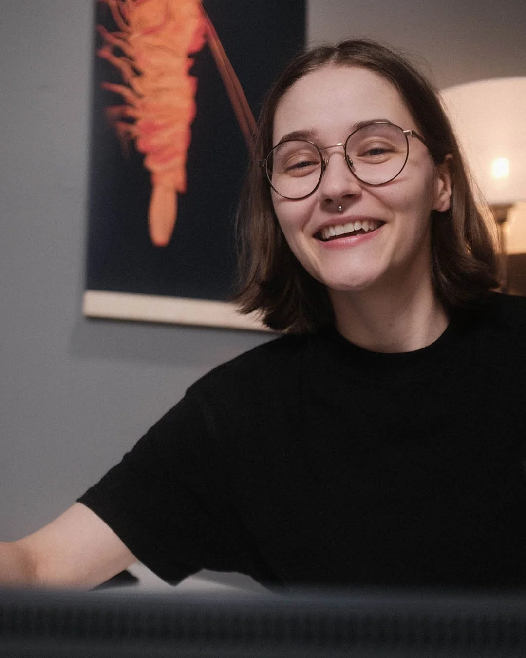 Lächelnde junge Frau mit Brille und Nasenpiercing, im Hintergrund ein Kunstwerk an der Wand, Lichtquelle rechts