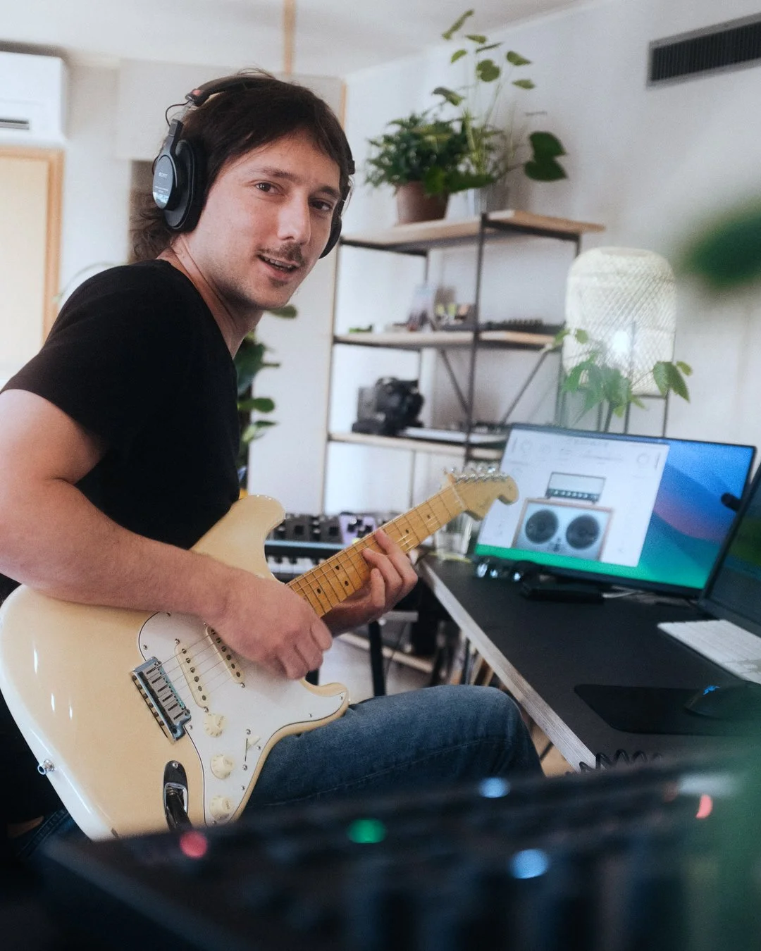 Junger Mann sitzt in einem Heimstudio, spielt Gitarre, trägt Kopfhörer, vor einem Computer und Musik-Equipment.