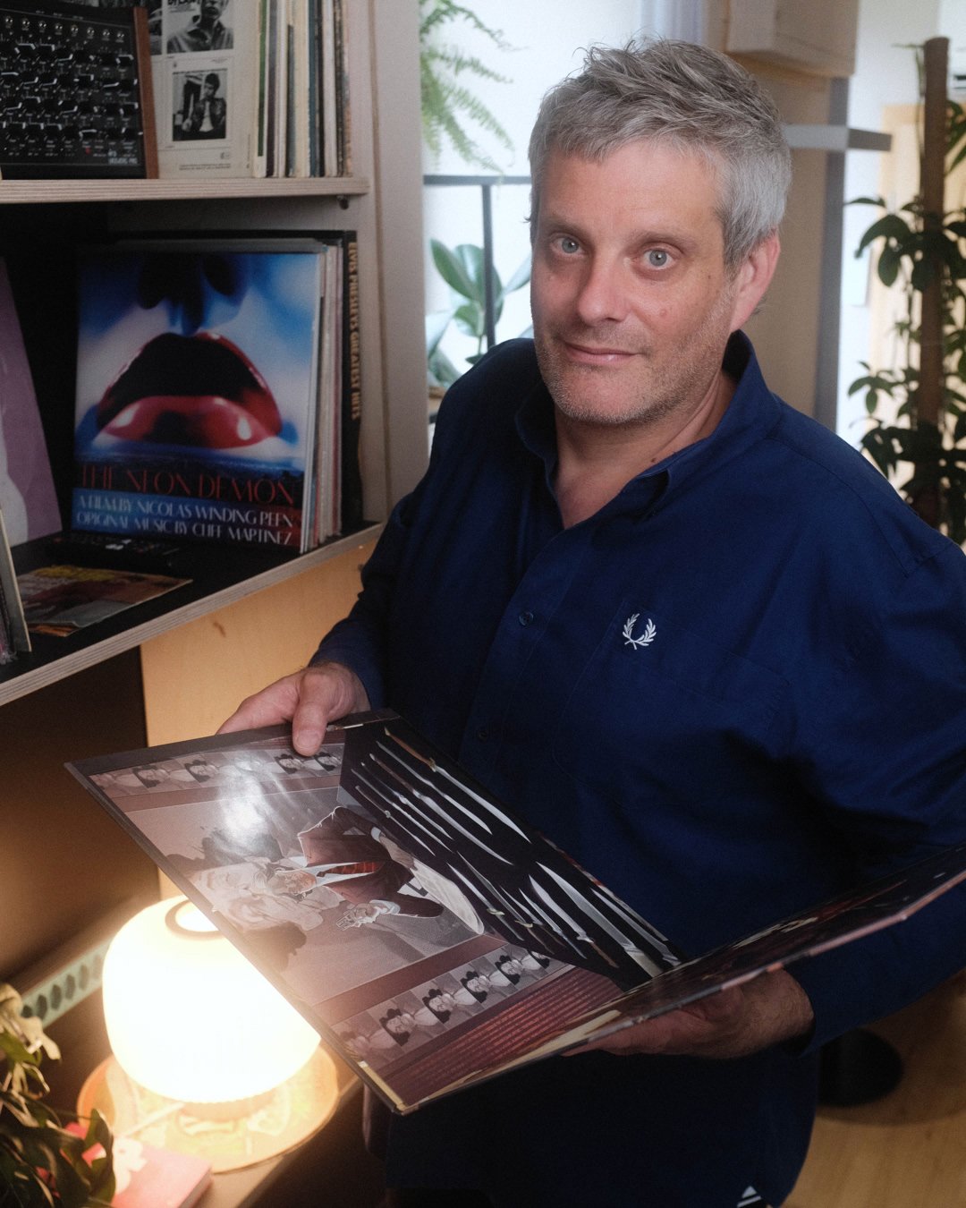 Ein Mann mit grauem Haar trägt ein dunkelblaues Hemd und hält ein Fotobuch, während er in einem Raum voller Bücher und Pflanzen steht.