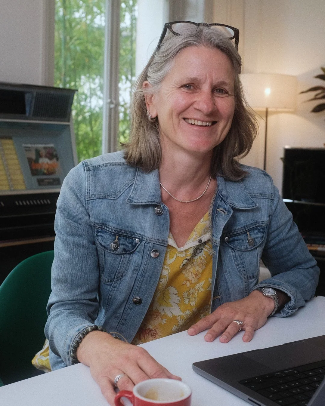 Eine lächelnde ältere Frau mit grauem Haar, Sonnenbrille auf dem Kopf, trägt eine Jeansjacke und eine gelbe Bluse mit Blumenmuster, sitzt an einem weißen Tisch in einem Raum mit einem Klavier im Hintergrund und hält eine Tasse Kaffee in der Hand.