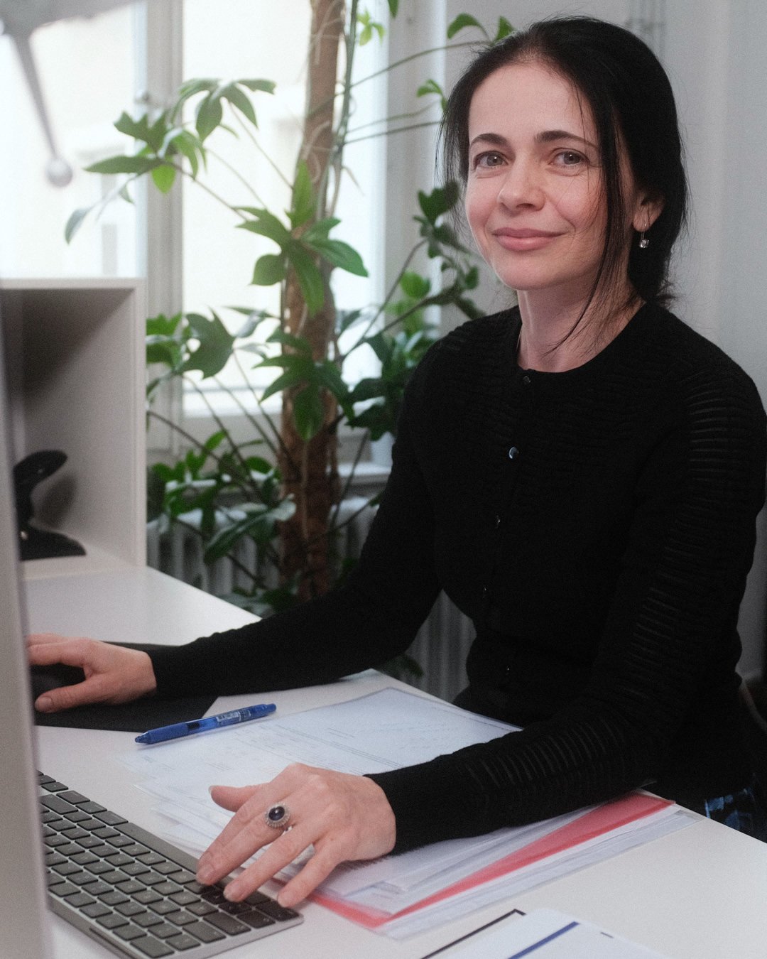 Eine Frau sitzt an einem Schreibtisch mit einem Laptop, einen Stift und Dokumenten, im Hintergrund sind Zimmerpflanzen und ein Fenster.