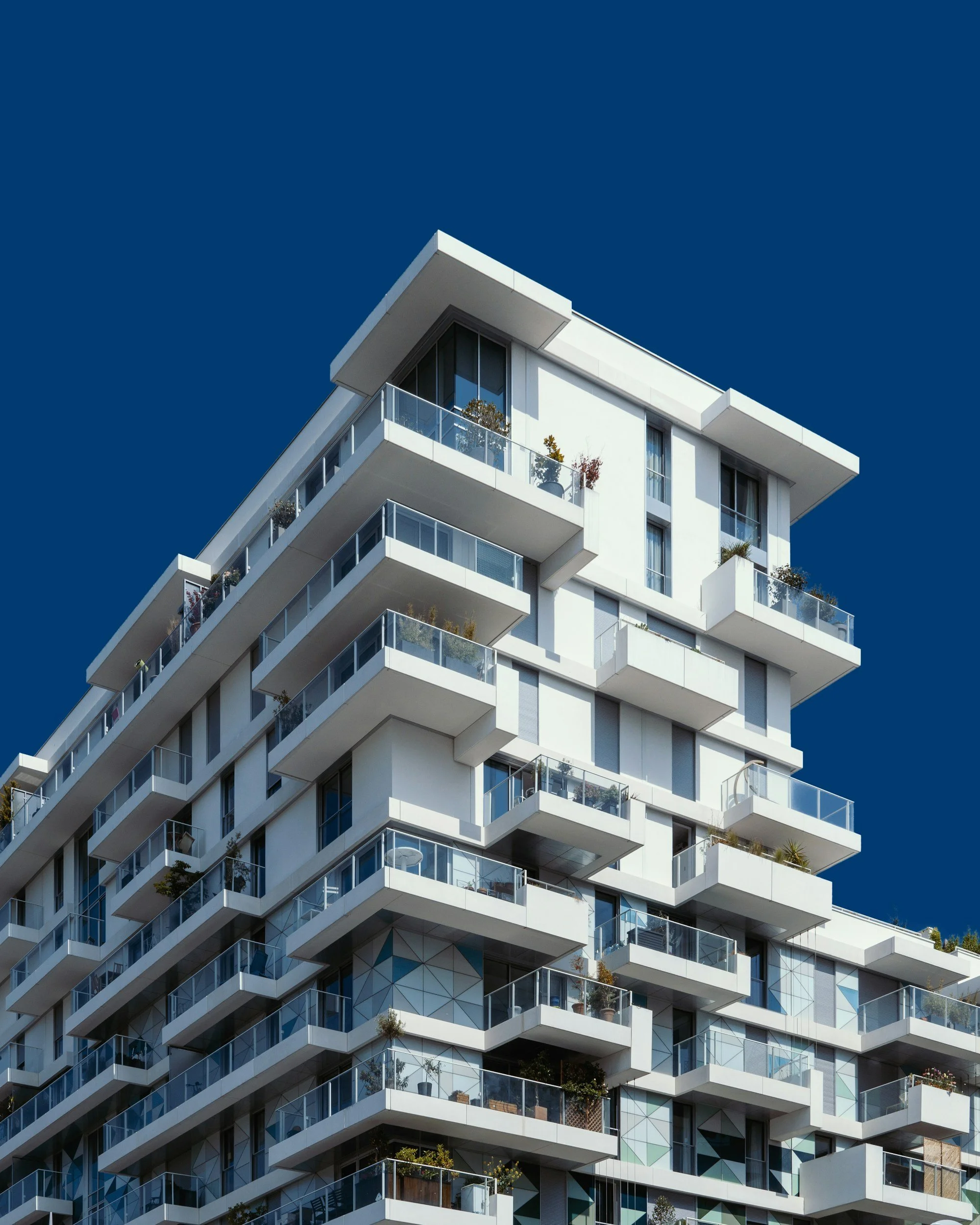 Modern multi-story residential building with white exterior walls, glass balconies, and various potted plants on balconies against a clear blue sky.