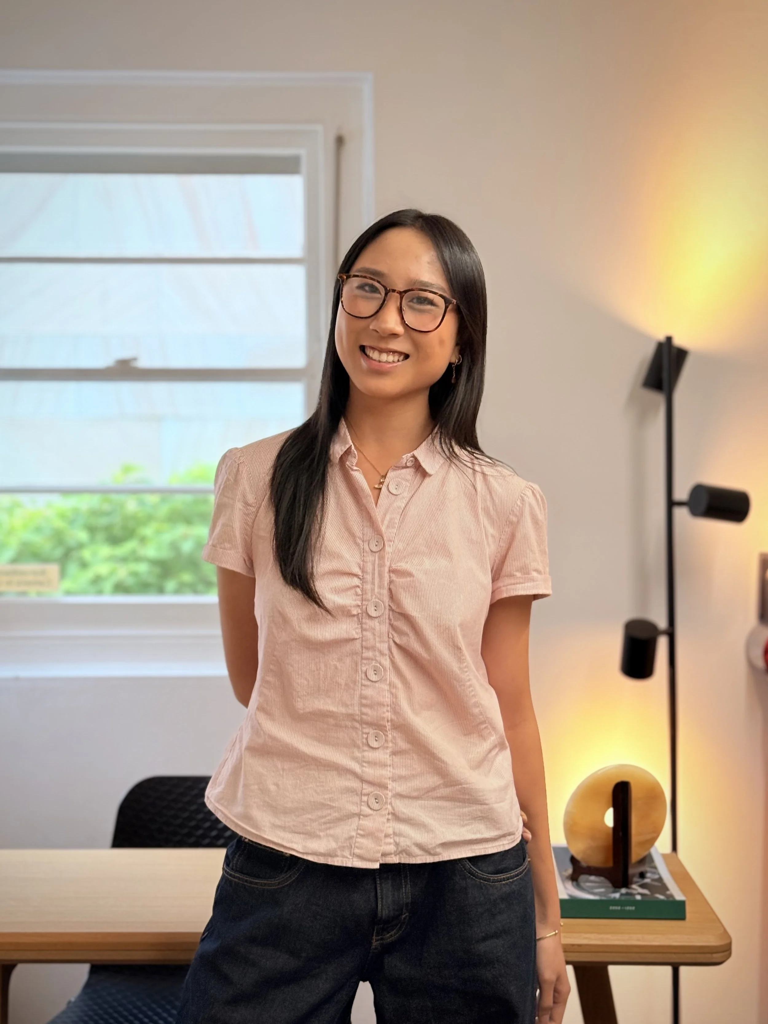 A woman with long black hair, glasses, and wearing a light pink blouse, stands inside a modern office with white walls, a window with greenery outside, and a floor lamp with yellow light in the background.
