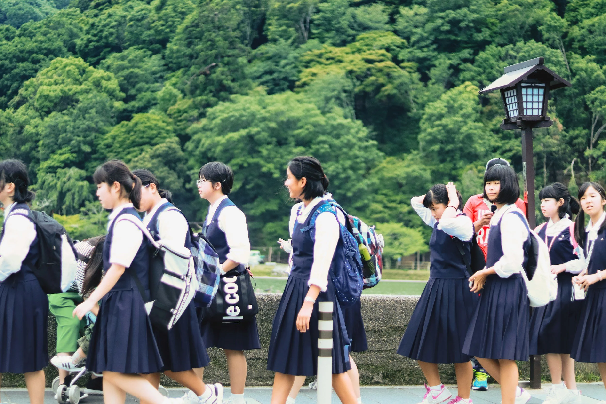 Group of Japanese school girls walking outdoors with lush green trees in the background, some wearing backpacks and uniforms.