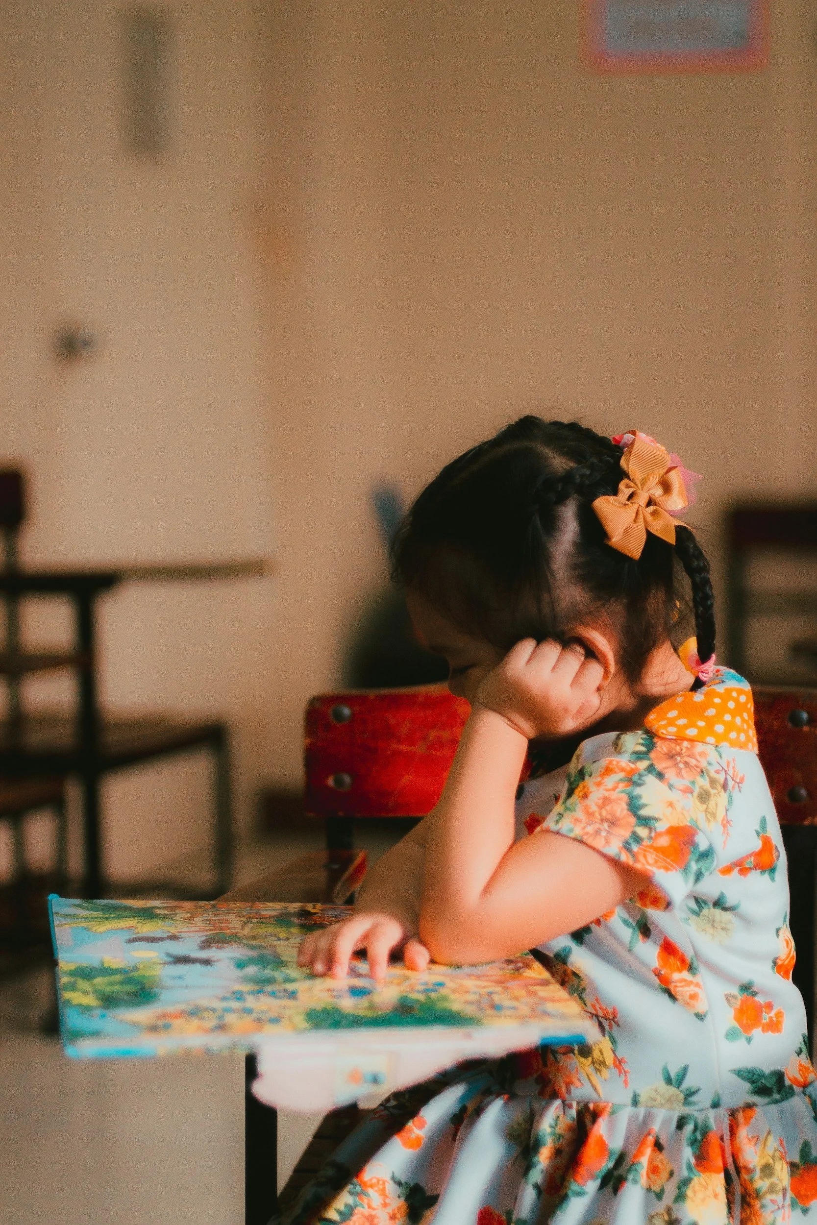 A young girl with dark hair in braids, wearing a floral dress with orange accents and peach-colored bows, is sitting at a table with her head resting on her arm, looking down at a puzzle on the table.