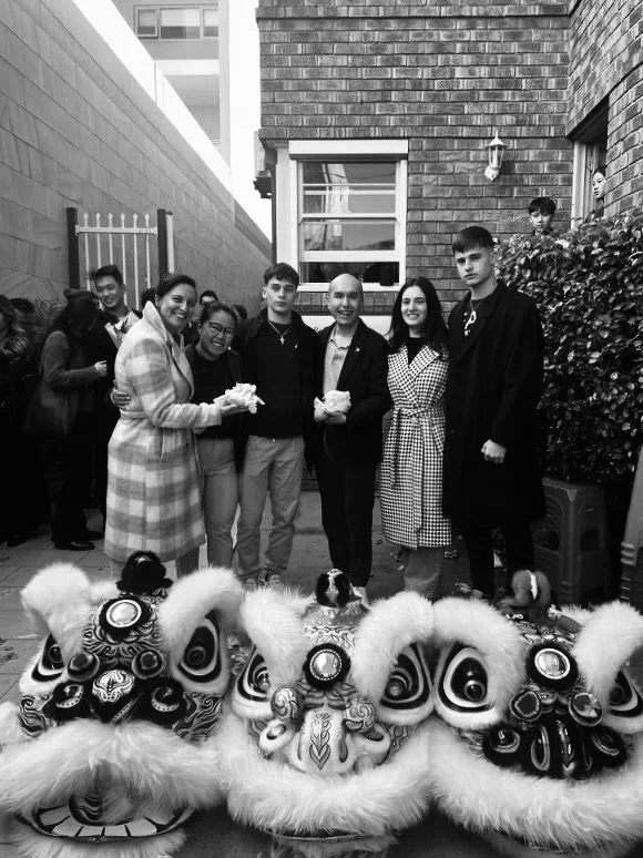 Group of people, including children and adults, gathered outdoors for a celebration. They are standing behind traditional Chinese lion dance costumes, which are decorated with intricate patterns and fluffy fur. Some people are holding small packages or envelopes, likely part of the festivities.