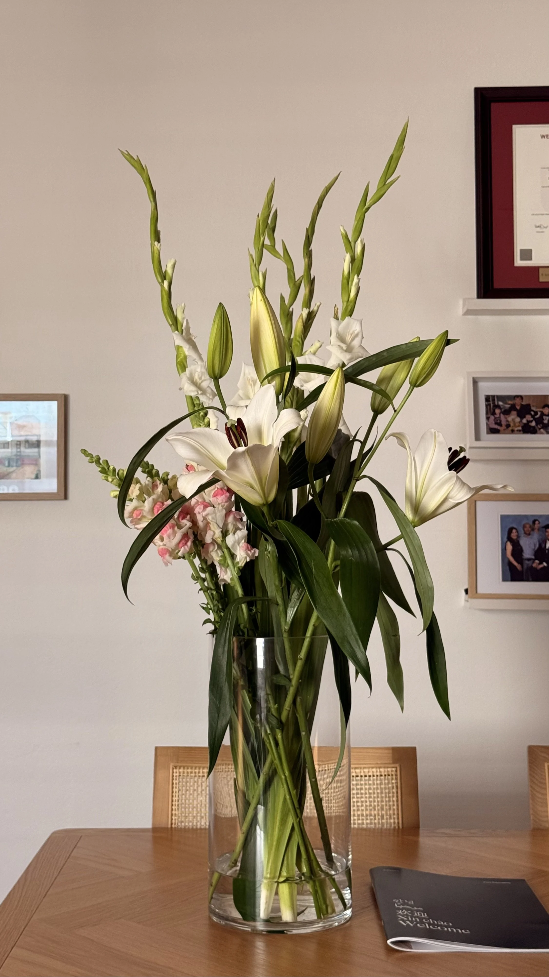 A clear glass vase with a large bouquet of white lilies and pink snapdragons on a wooden table.