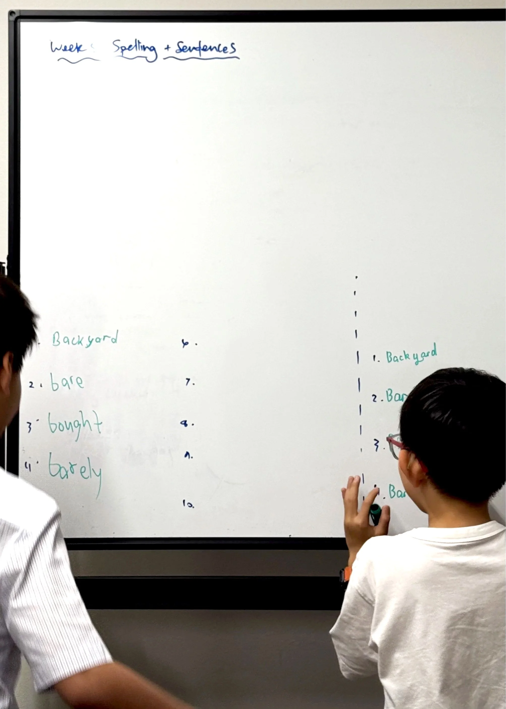 Two children standing in front of a whiteboard with handwritten list of words and sentences related to spelling and sentences, with one child writing on the board.