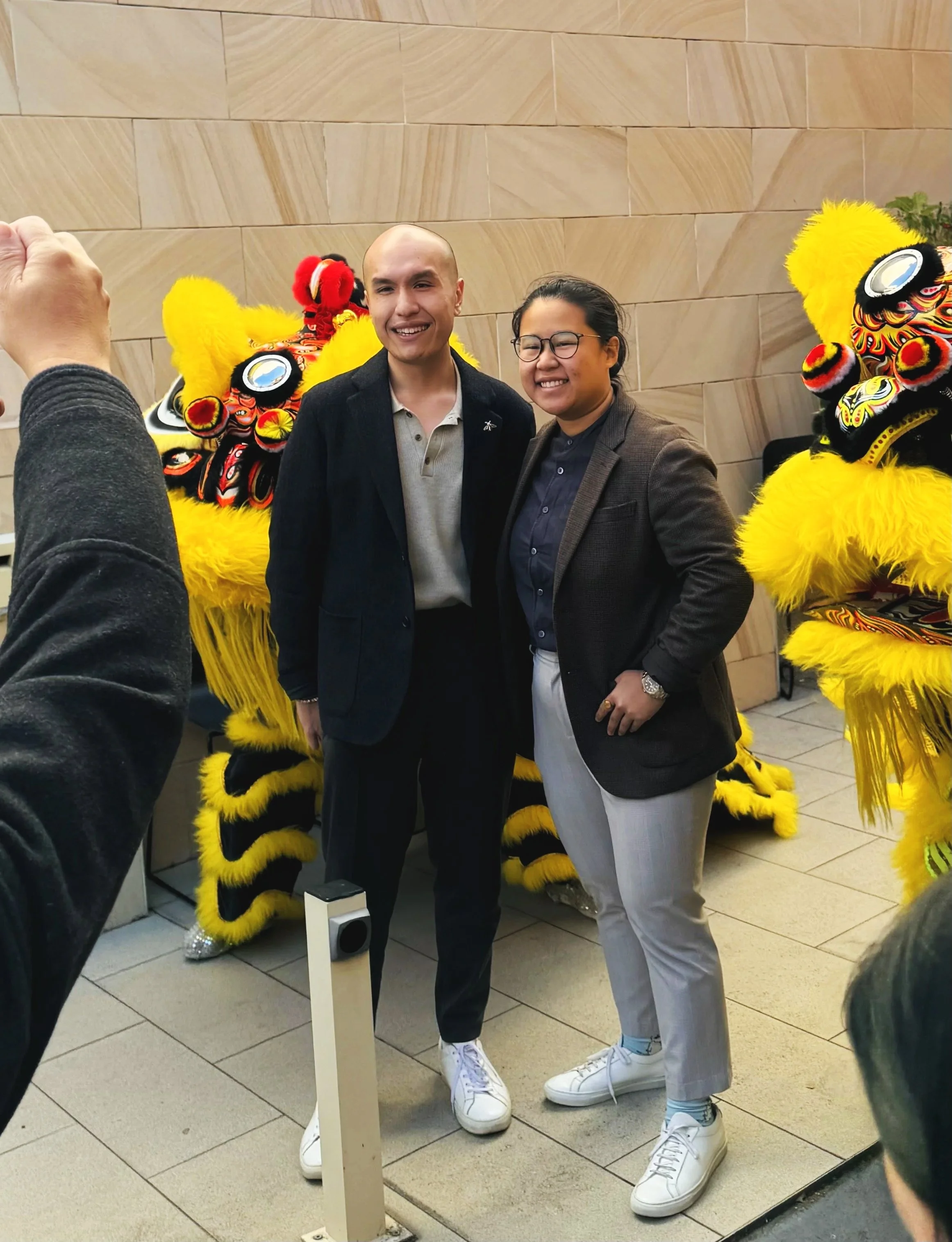 Two people posing for a photo with two lion dance costumes in the background. One is a man with a bald head wearing a dark blazer and light-colored shirt, and the other is a woman with glasses and black hair tied back, wearing a dark blazer and light-colored pants. They are standing close together, smiling at the camera.