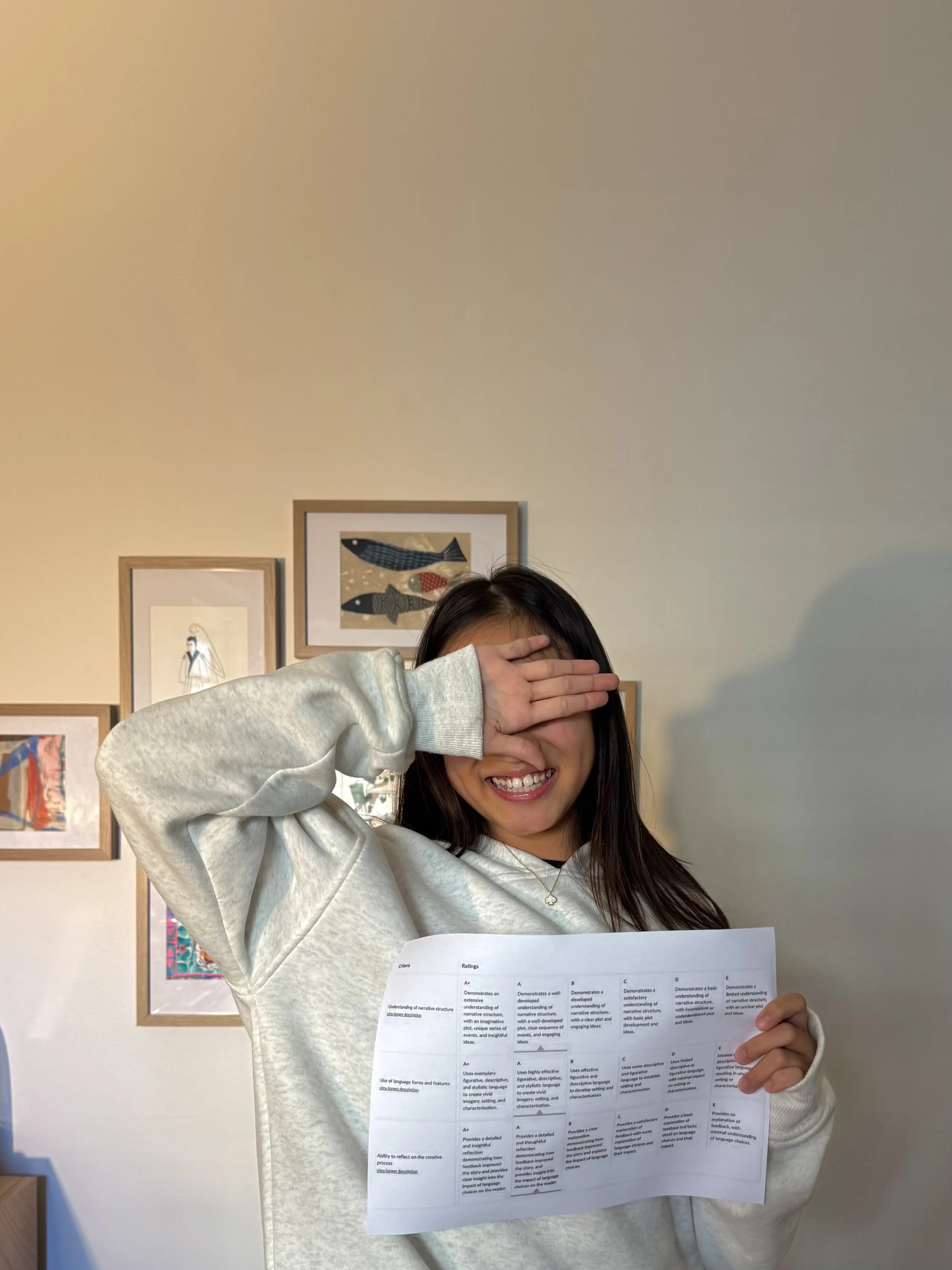 A young woman with long dark hair smiling and covering her eyes with her hand, holding a sheet of paper in a room with framed artwork on the wall behind her.