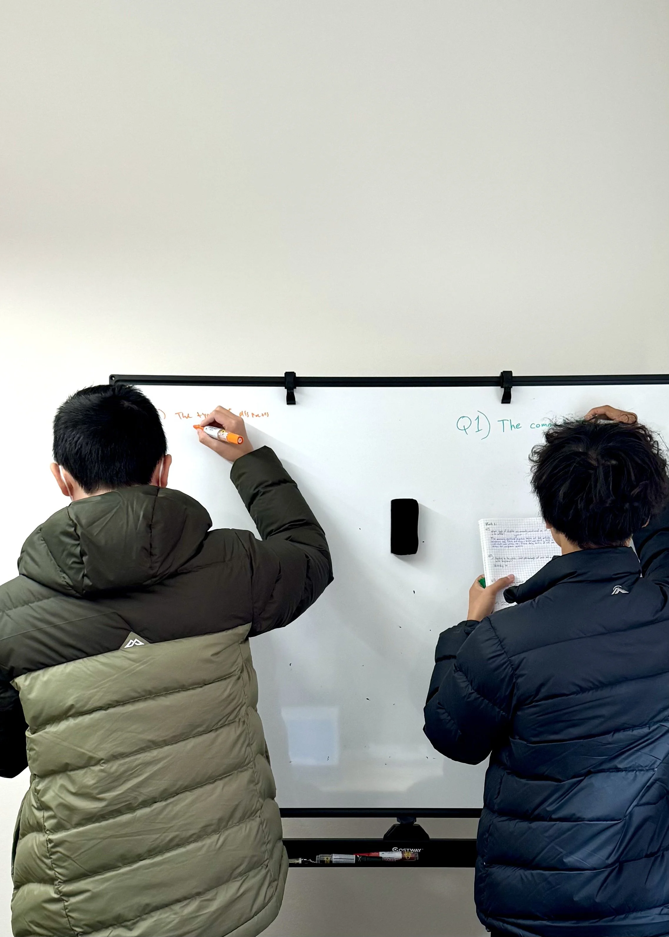 Two people wearing winter jackets standing and writing on a whiteboard in a classroom.