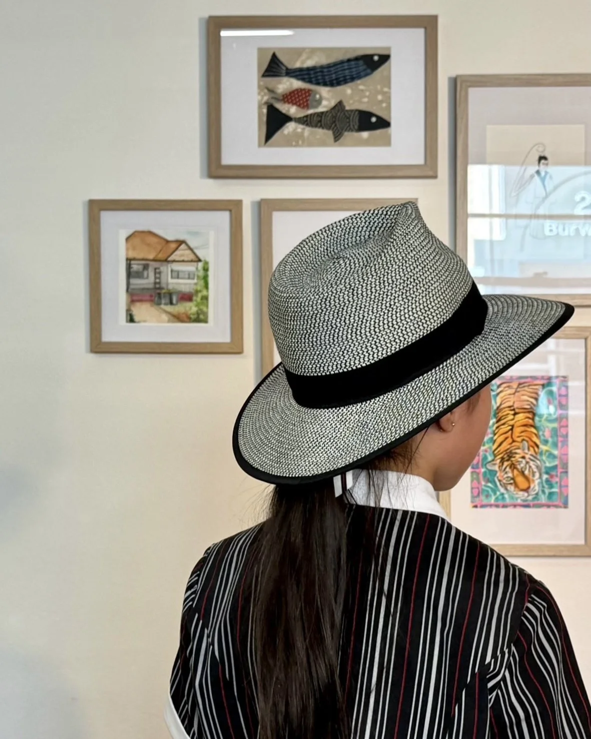 A woman with long dark hair wearing a wide-brimmed straw hat with a black band, viewed from behind, standing in an art gallery with framed artwork on the wall.