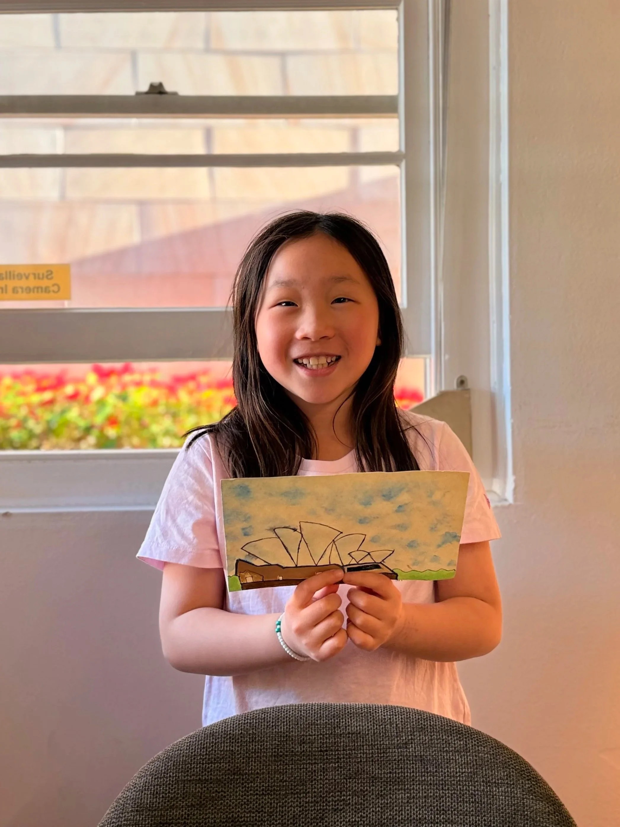 A young girl with dark hair smiling while holding a colorful drawing of a landmark with a blue sky, clouds, and green grass.