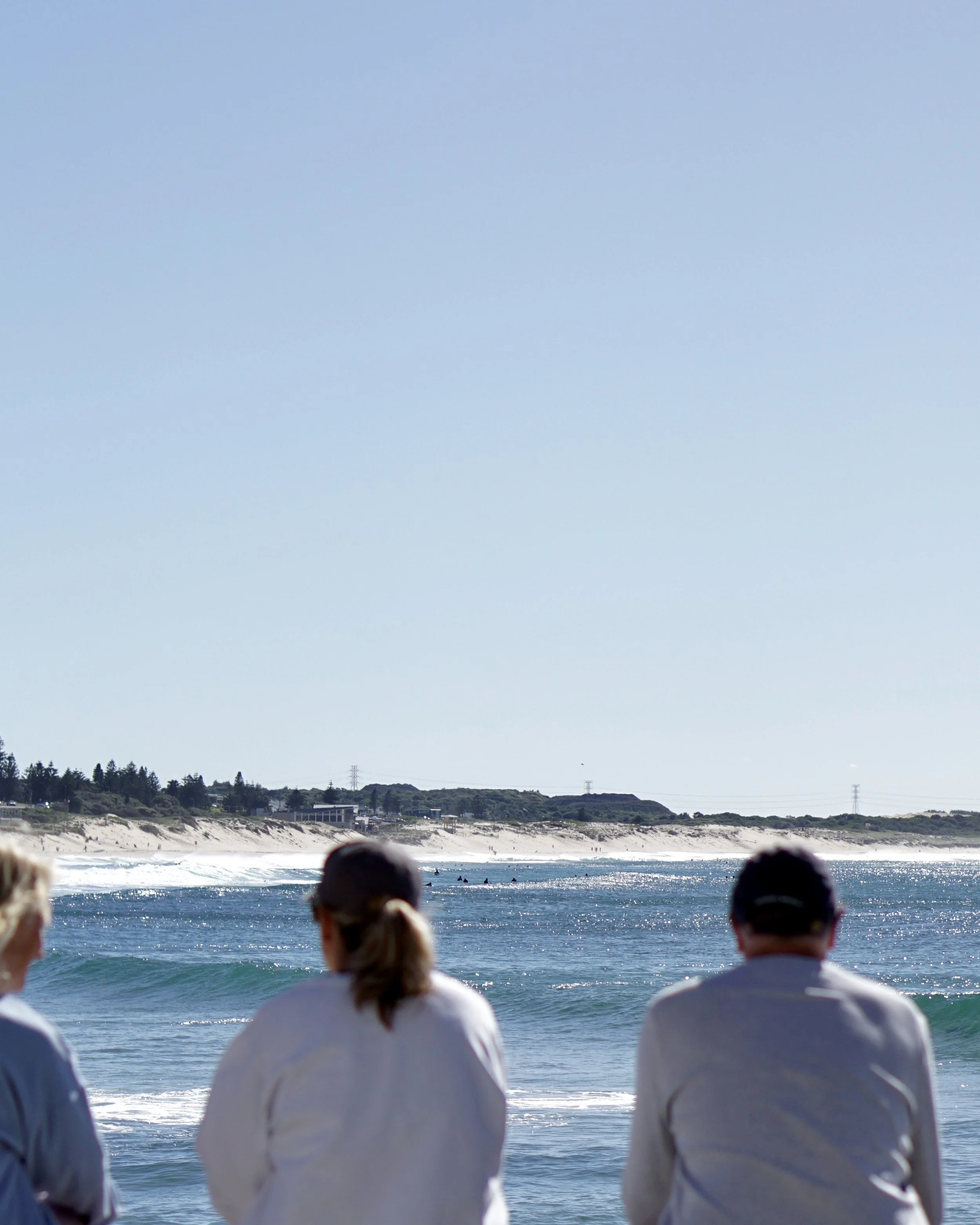 Three people wearing white jackets sitting on a beach, facing the ocean with small waves and a sandy shoreline in the background under a clear blue sky.