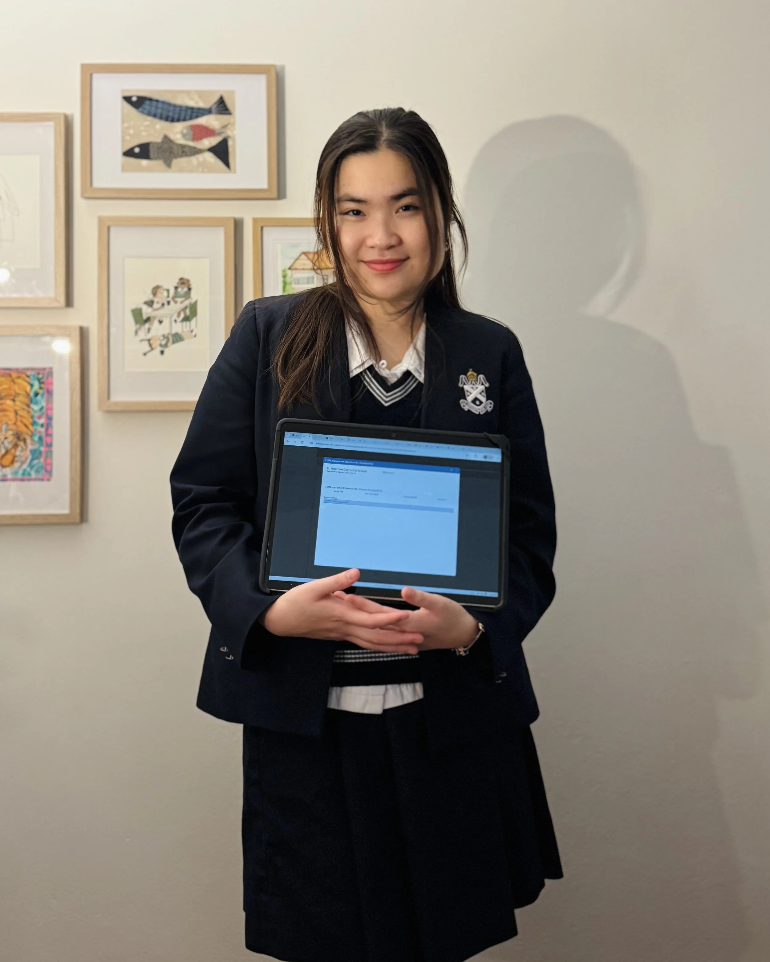 Young woman in school uniform holding a tablet, standing in front of a wall decorated with framed artwork.