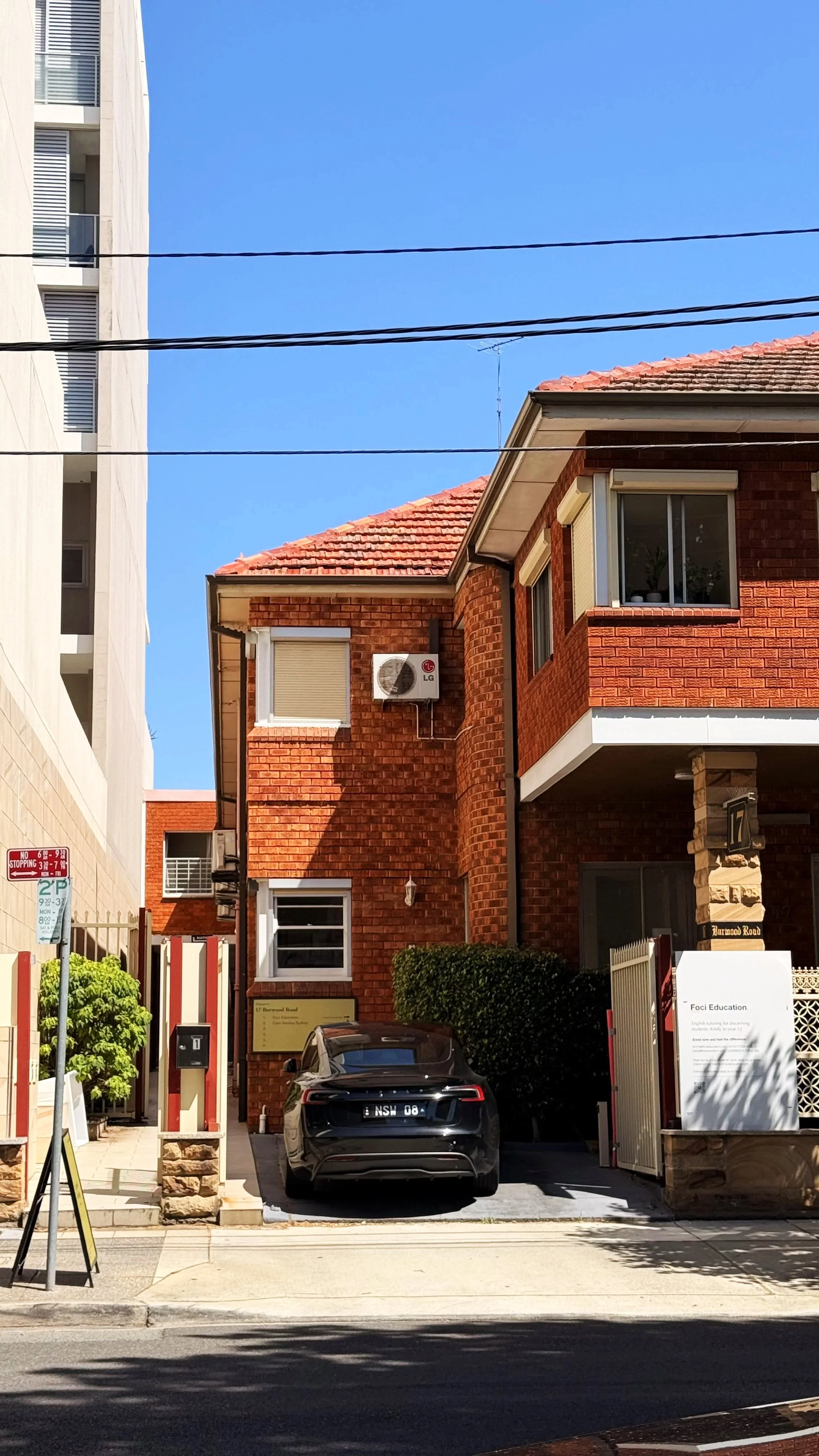 A black car parked in front of a red-brick building with a small hedge and a white sign. The building has an air conditioning unit on the upper wall and a window with blinds. There are black power lines overhead and a clear blue sky.