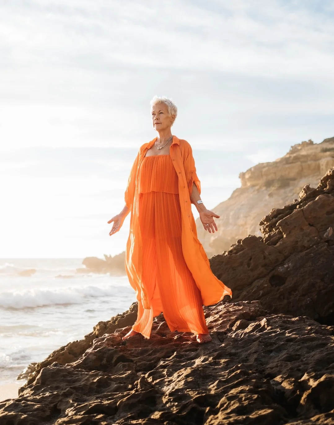 An elderly woman in an orange flowing dress and matching open shirt standing barefoot on rocky shore at the beach with cliffs in the background during sunset.
