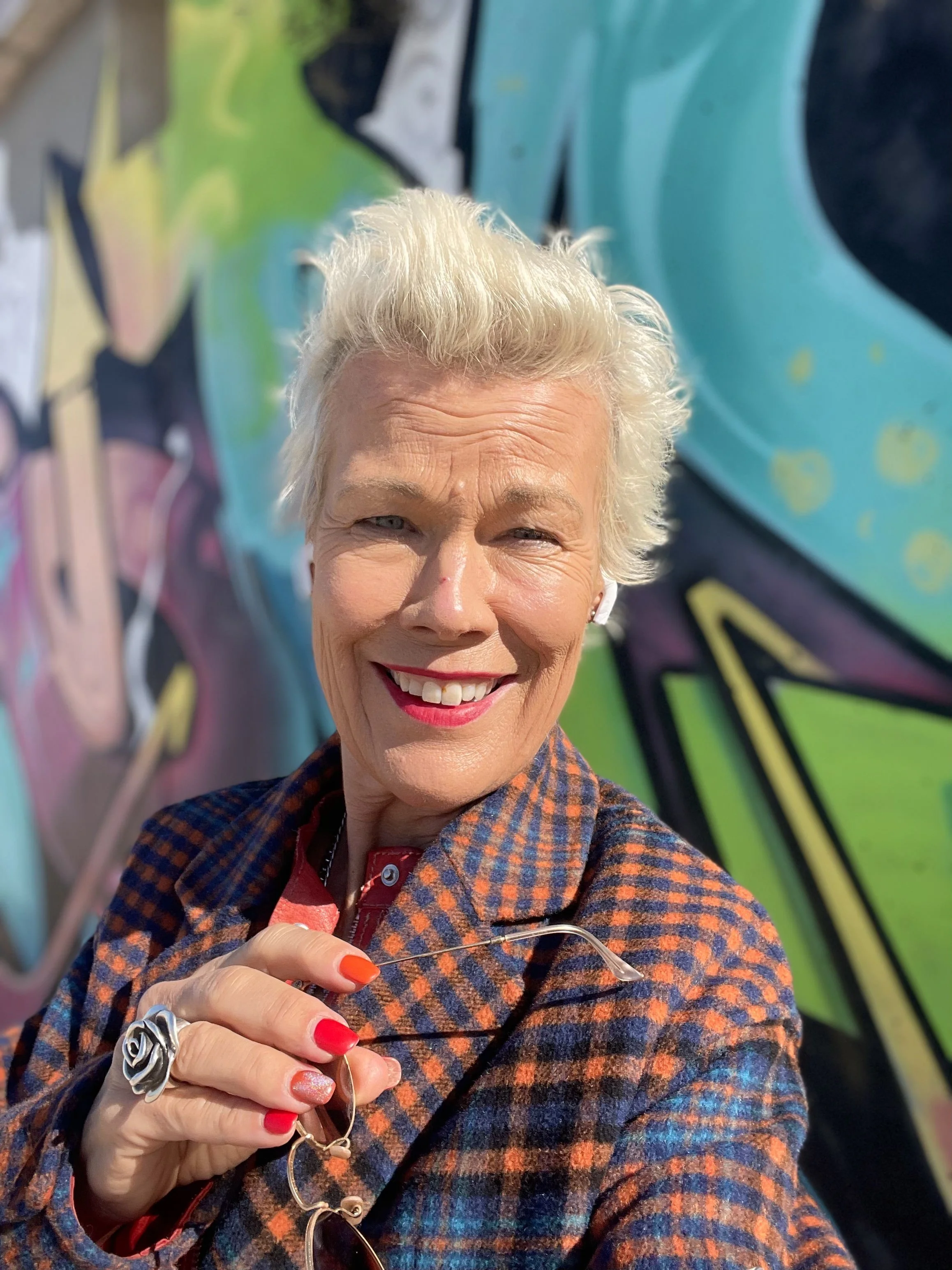 A smiling woman with short, platinum blonde hair, wearing a checkered blazer and holding sunglasses in front of a colorful graffiti wall.