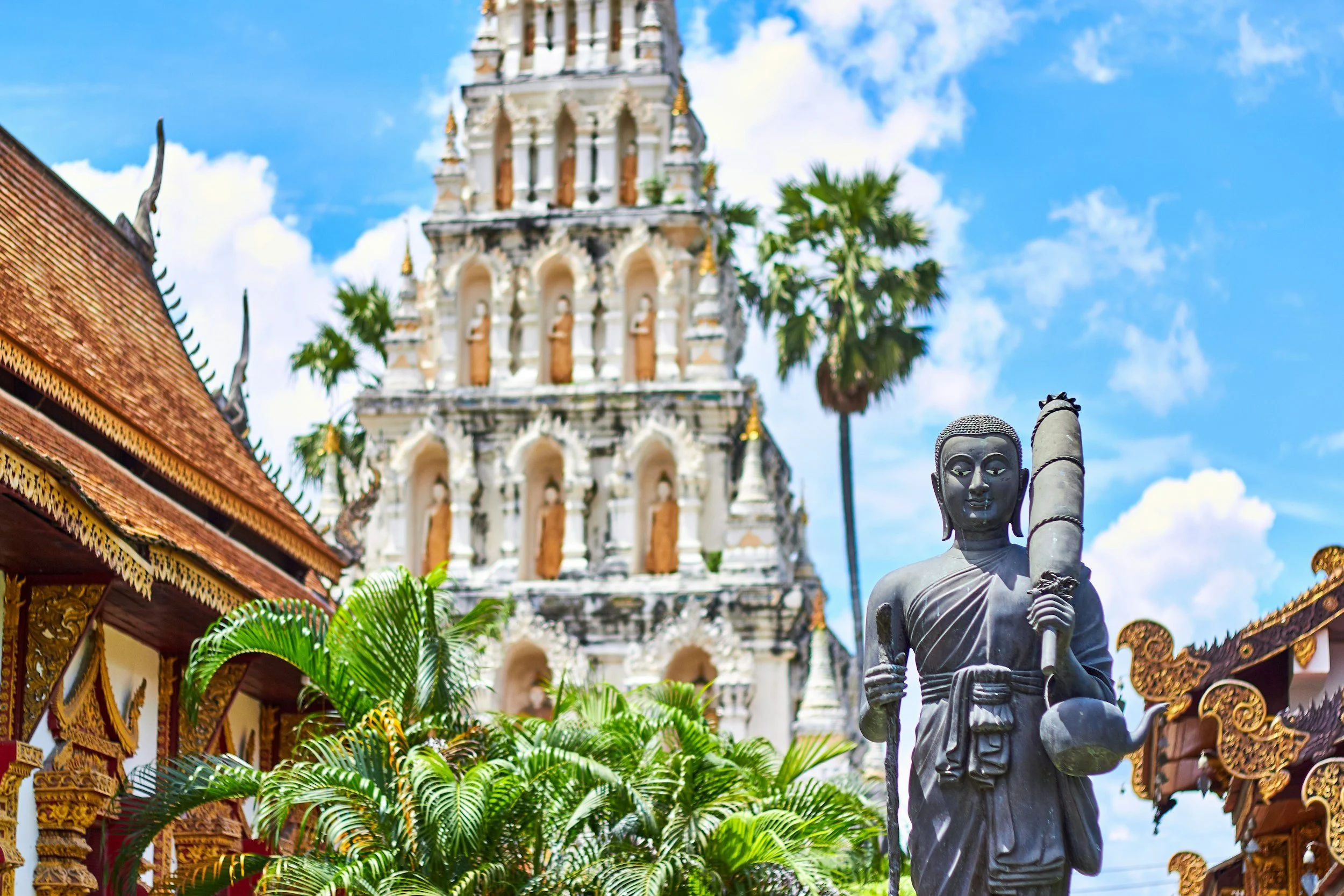 A decorative sculpture of a person holding a staff in front of a temple with ornate architecture, surrounded by green palm trees and a blue sky with clouds.
