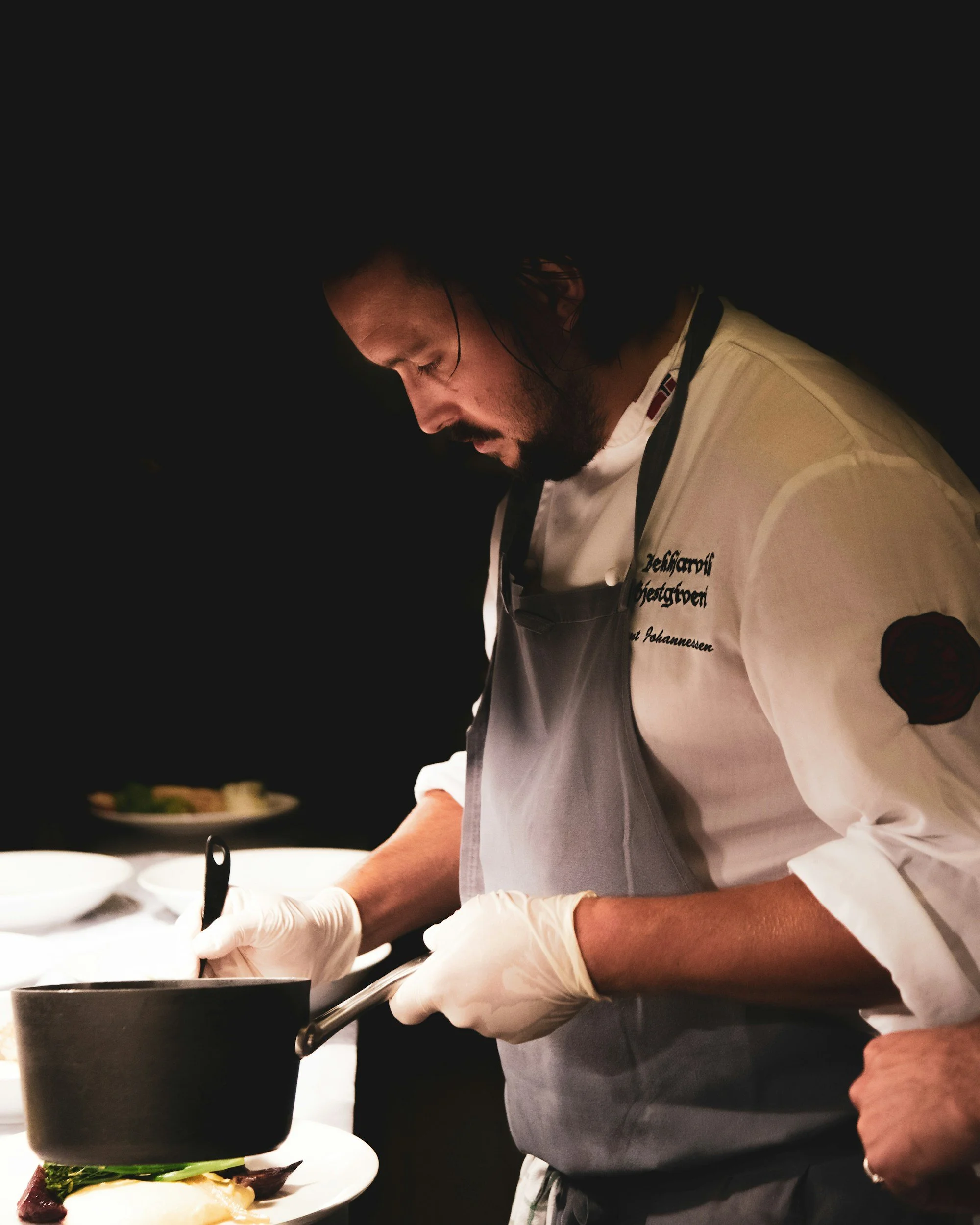 A male chef wearing a white apron and gloves preparing food in a kitchen, with a plate of food in front of him.