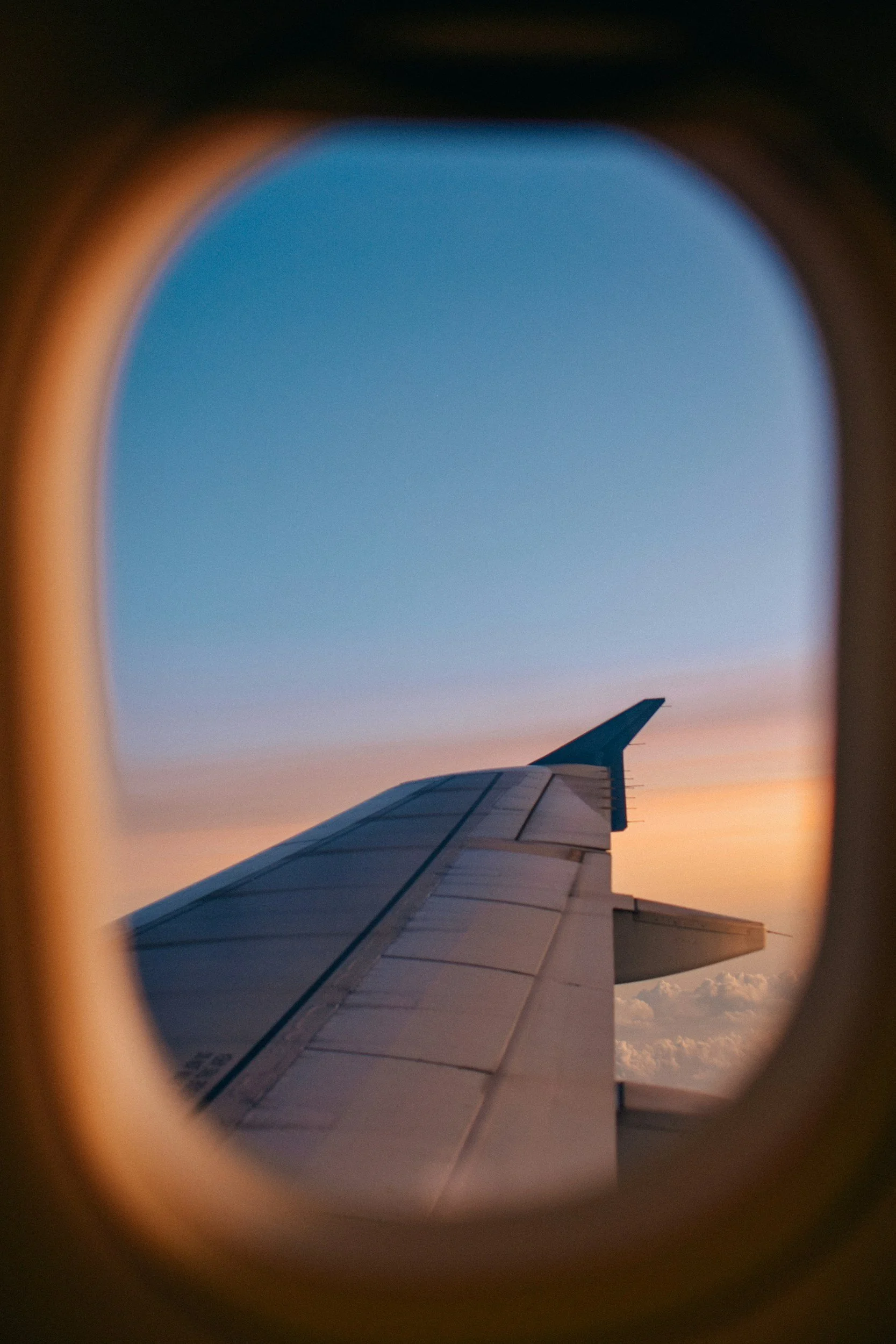View of an airplane wing through an airplane window with a sunset sky and clouds in the background.