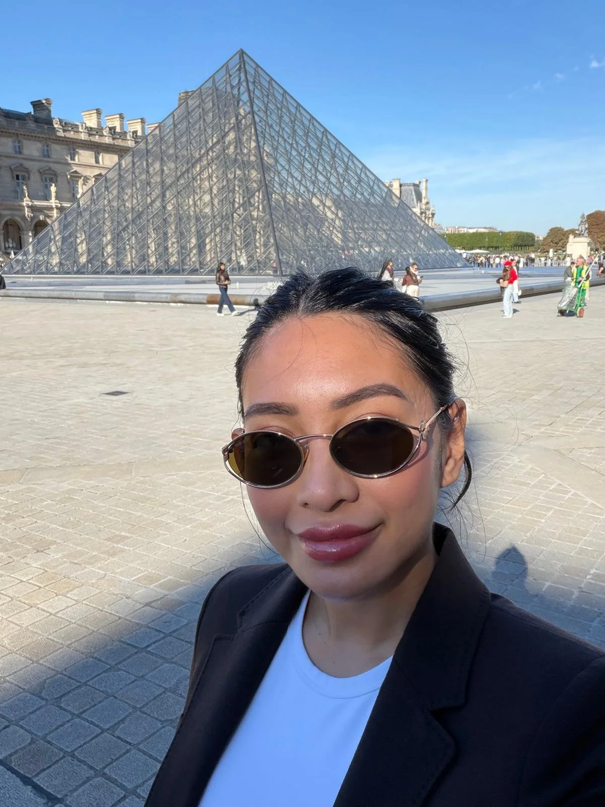 A woman with dark hair and round sunglasses taking a selfie in front of the Louvre Pyramid in Paris, France on a clear day.