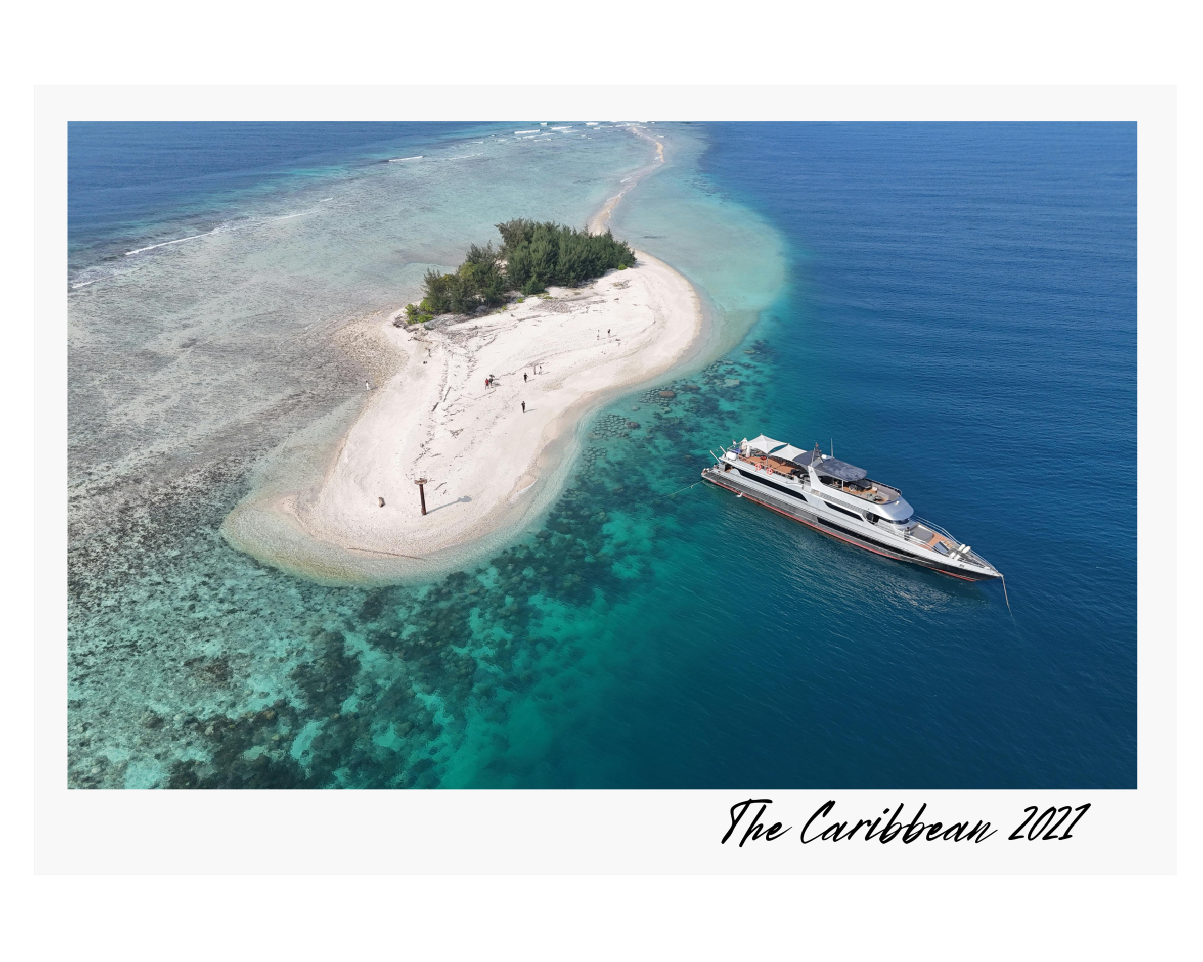 Aerial view of a small, sandy island with green trees in turquoise waters, with a yacht anchored nearby, in the Caribbean in 2021.