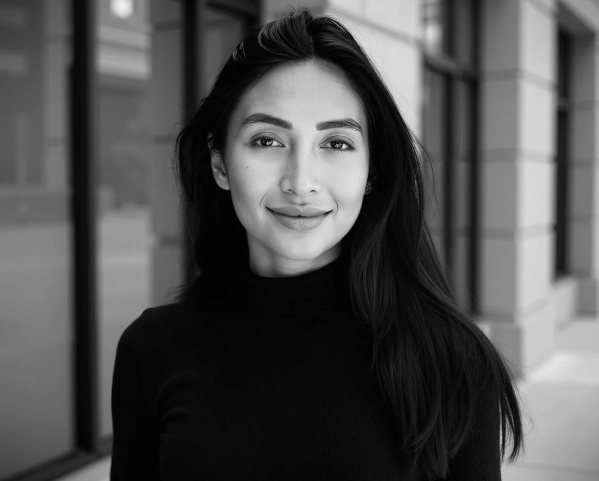 A black-and-white portrait of a woman with long dark hair, wearing a turtleneck sweater, standing outdoors in front of a modern building with glass and stone architecture.