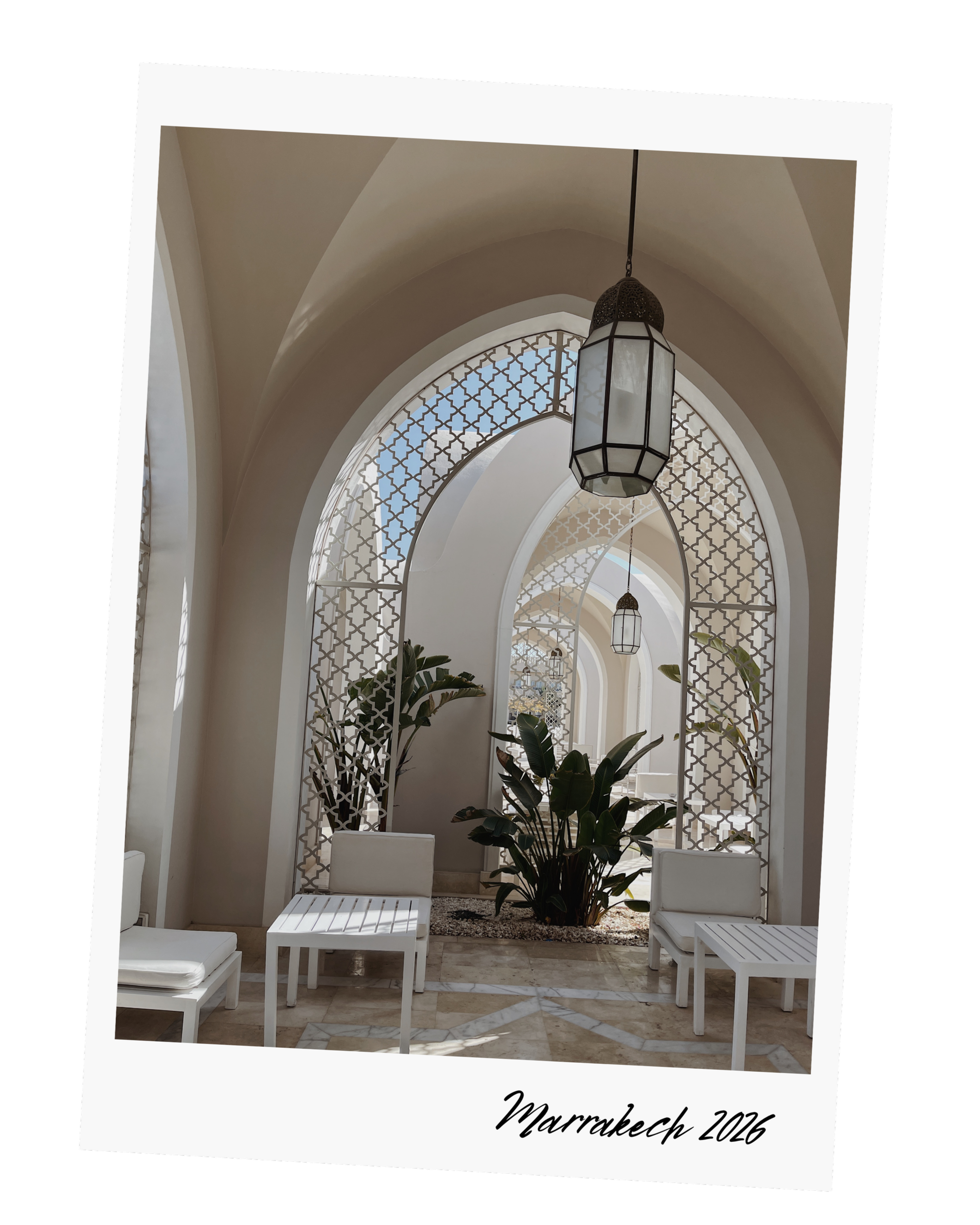 Photo of an arched corridor with white furniture and a potted plant, featuring decorative lattice windows and hanging lamps, with "Marrakech 2026" written at the bottom.