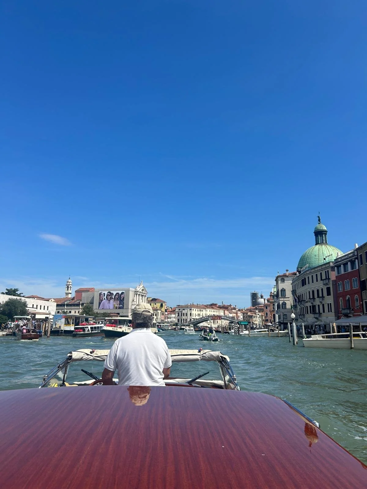A man steering a boat on a canal in Venice, Italy, with historic buildings and a green-domed church in the background under a clear blue sky.