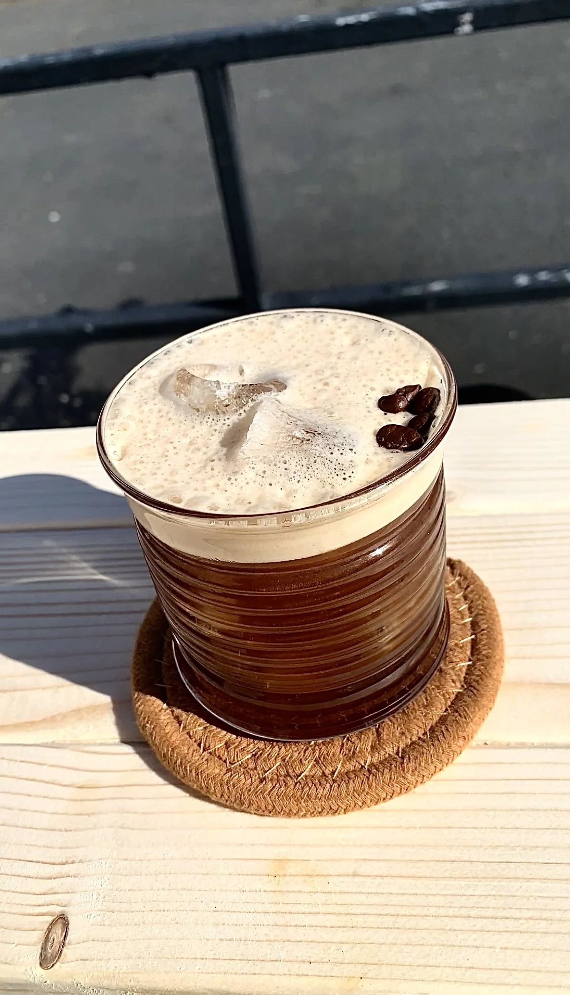 A glass of iced coffee topped with foam and coffee beans, placed on a round cork coaster on a light wooden surface outdoors.